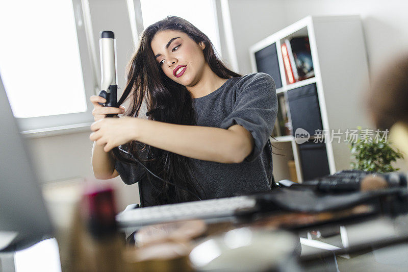 坐在梳妆台上看着电脑和卷发棒的女人。在冠状病毒大流行期间，一名女子在网上看教程做头发。