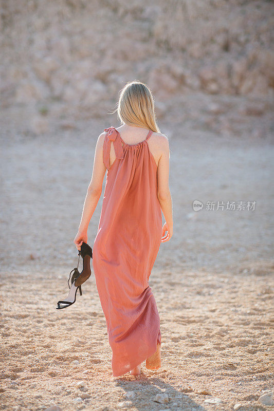 后视图，美丽的女人穿着夏装，手拿凉鞋走在石头沙漠