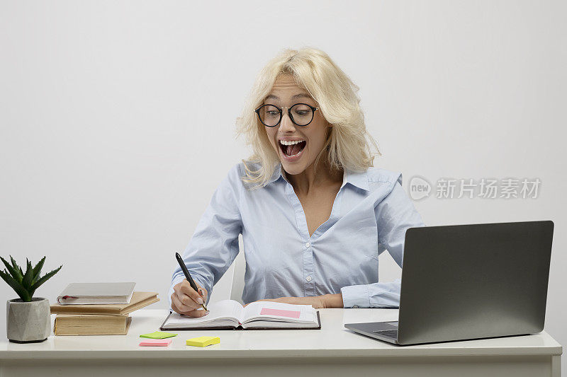 惊讶的女商人在笔记本电脑上工作，在笔记本上做笔记，惊讶地阅读她的笔记在工作场所