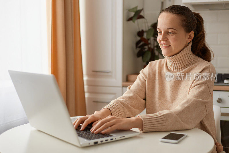 黑色头发的女人，穿着米色毛衣，坐在厨房里，用笔记本电脑工作，敲击键盘，表达积极的情绪。