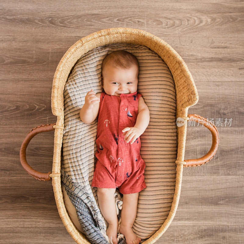 一个18周大的男婴穿着红色的航海服躺在一个舒适的奶油色条纹棉毯在海草摩西篮子