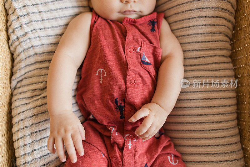一个18周大的男孩的嘴唇和手穿着红色航海服躺在一个舒适的奶油色条纹棉毯在海草摩西篮子