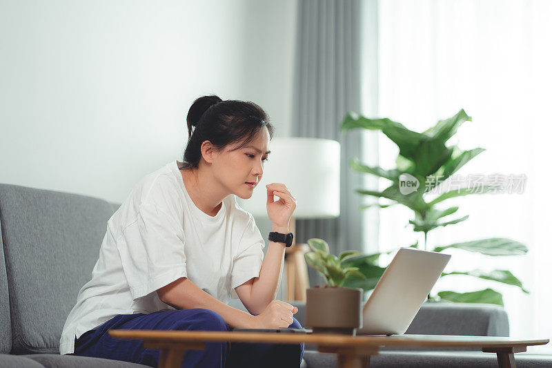 一名亚洲妇女坐在家里的沙发上，用笔记本电脑工作，发烧了。