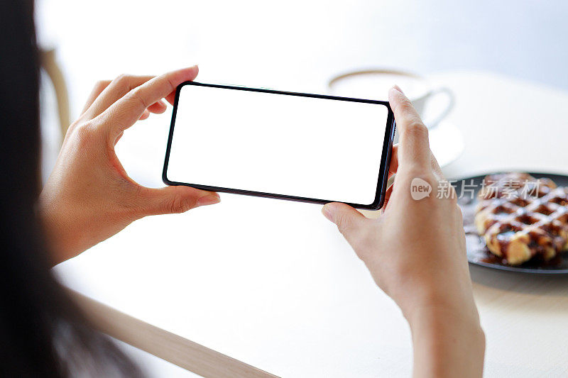 一名女子的手拿着空白屏幕的智能手机，在咖啡馆里为设计模型拍照。