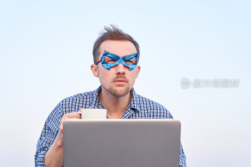 戴着蓝色面具的超级英雄IT男在户外拿着咖啡杯拿着笔记本电脑