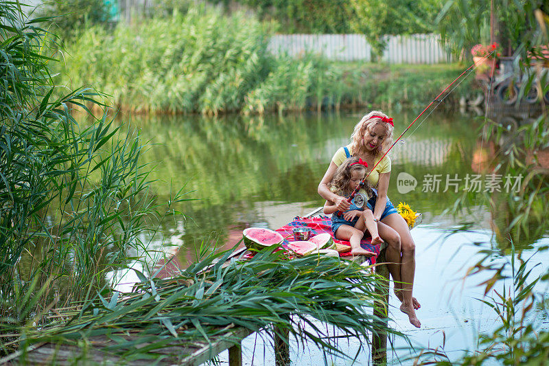 金发碧眼的母亲和三岁的女儿穿着牛仔短裤，系着红色蝴蝶结的腰带，在河上的码头上吃西瓜和鱼