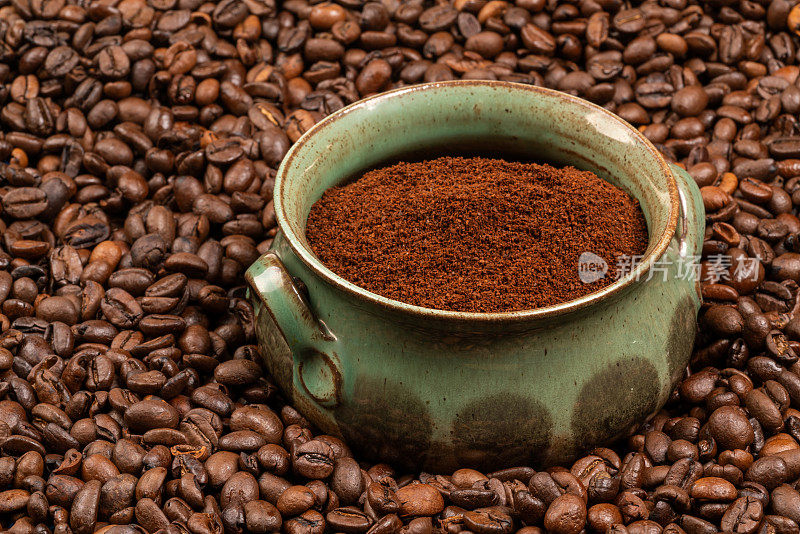 一个绿色的陶制杯子，在许多咖啡豆中间放着咖啡粉。多样性。口味不同。快乐。觉醒。滋补饮料。咖啡的香味。早餐。选择。每日能量