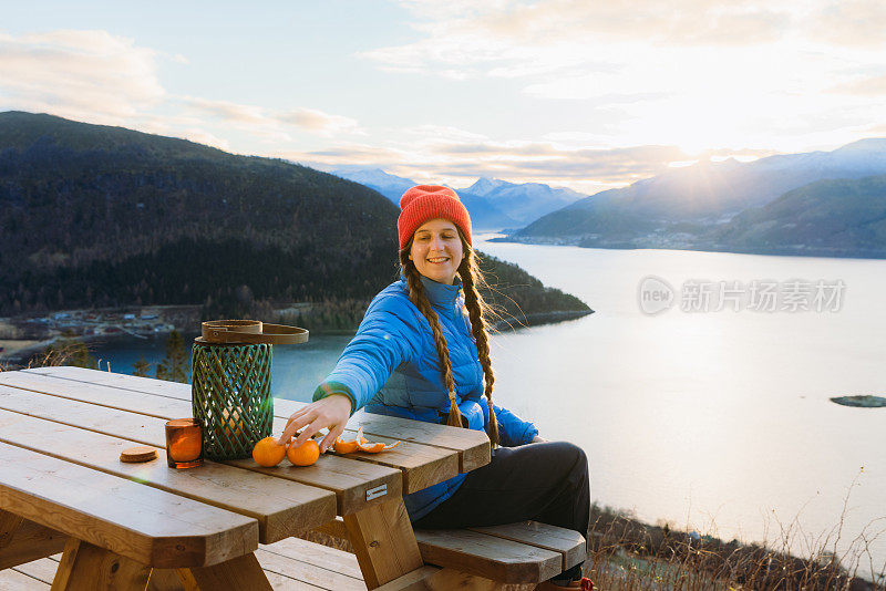 在挪威的日落时分，戴着红帽的快乐女人一边吃橘子，一边欣赏峡湾的美景