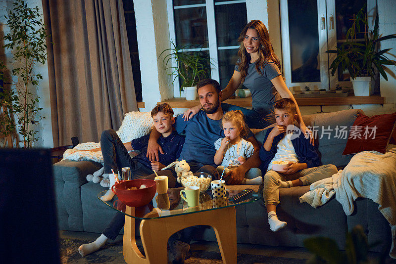幸福美满的家庭，晚上父母和孩子坐在家里的沙发上看电视，吃零食。放松