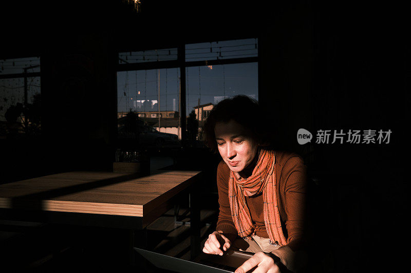 一个开朗的成熟女人坐在昏暗的咖啡馆里，用笔记本电脑和别人聊天