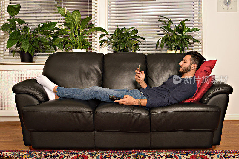 男人在沙发上休息，使用手机和电视遥控器