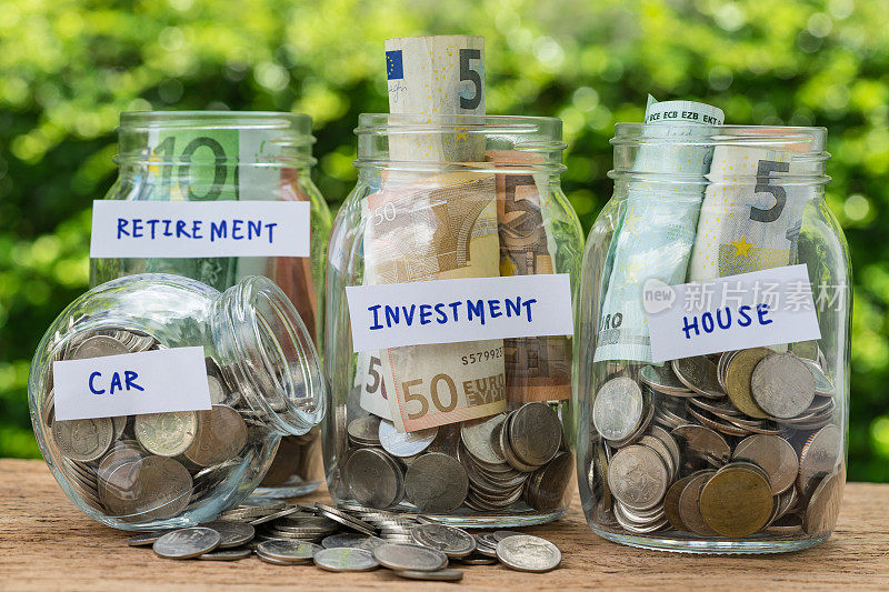 一组装满硬币和钞票的玻璃瓶瓶子贴上了投资，房子，汽车和退休作为储蓄或投资概念