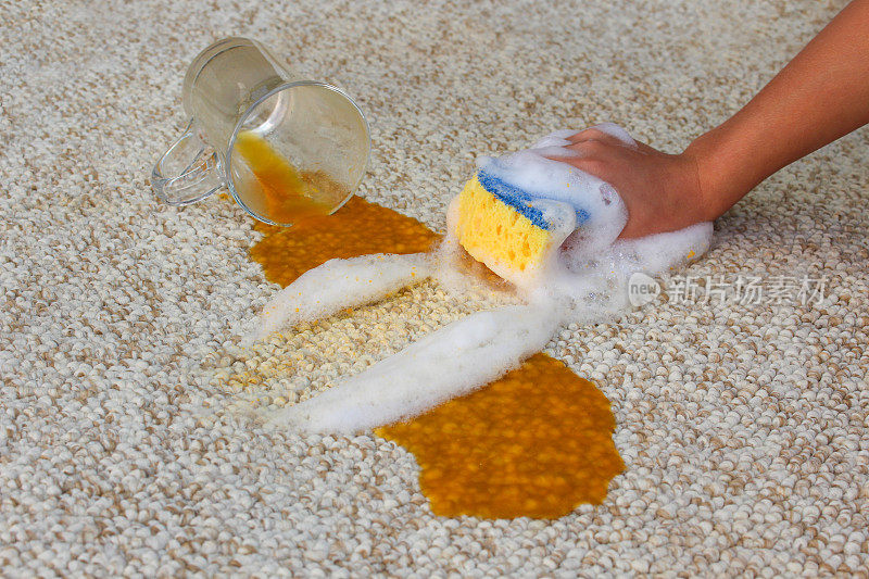 一杯果汁掉在地上，洒了一地。女人的手用海绵和洗涤剂清洗地毯。