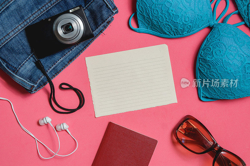 空白核对表，护照，泳衣，牛仔裤，太阳镜，粉红色背景上的相机。顶视图旅游概念。