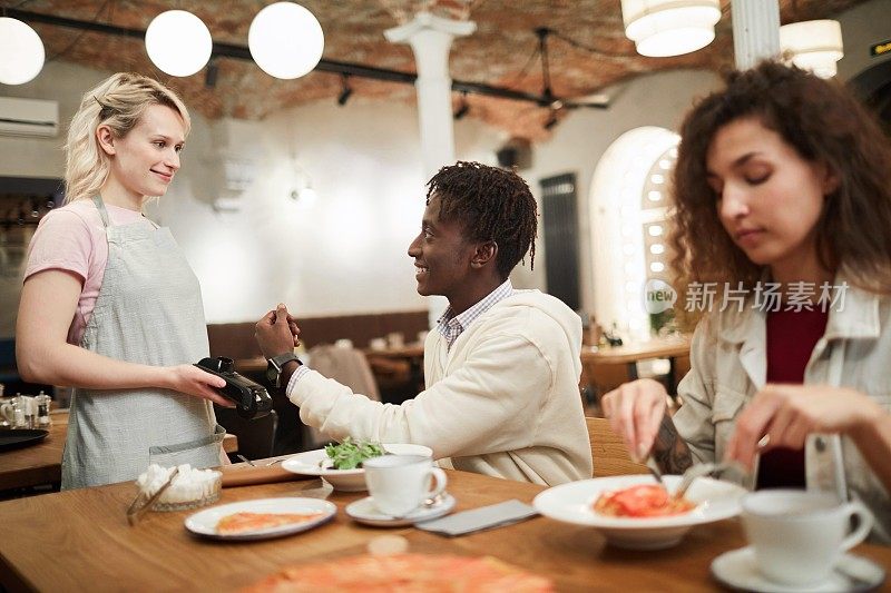 快乐兴奋的年轻非洲裔美国人坐在桌子上与女朋友吃意大利面，拿着智能手表在终端，而支付晚餐在咖啡馆