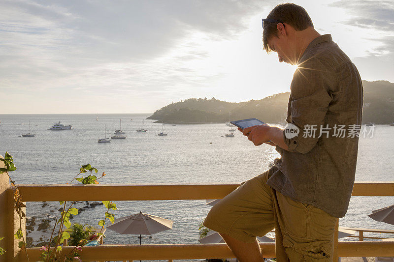 一个年轻人在一个平静的海湾上方的阳台上使用数码平板电脑