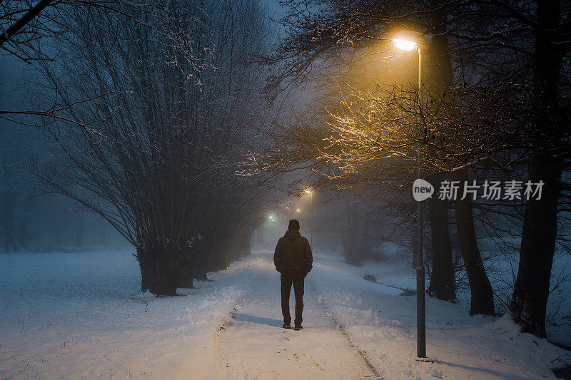 在雾中，一个年轻人独自走在黄色路灯下的人行道上。多雾的空气。雪夜的宁静气氛。后视图。