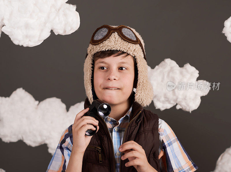 一个男孩玩着双筒望远镜，梦想着成为一名飞行员，穿着复古风格的夹克，戴着戴眼镜的头盔，棉絮缭绕，灰色的背景，略带棕色