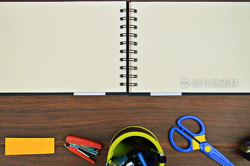 奶油色的空白页的开放式螺旋记事本，办公用品，如剪刀，订书机在一个木制的深棕色背景。