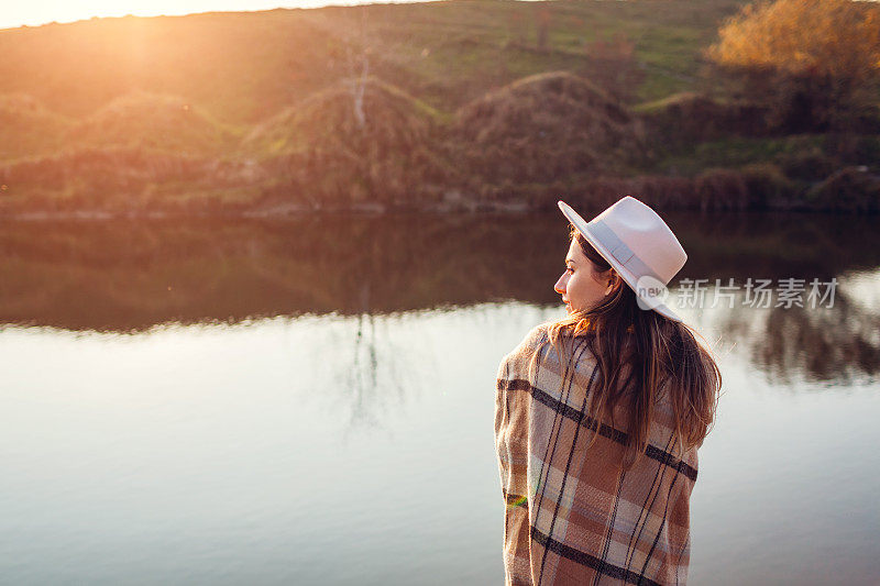 后视图的女人放松在秋天的河流在日落。戴着帽子的时尚女孩静静地享受着秋天的风景