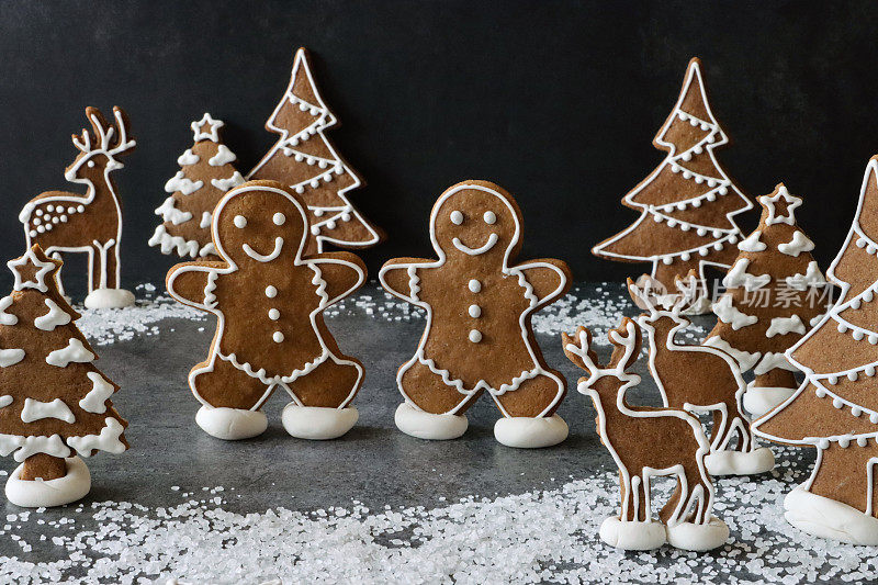 两个自制的姜饼人在白雪覆盖的池塘上滑冰，有驯鹿和树木的圣诞森林夜晚场景，用白色皇家糖霜装饰的圣诞主题饼干，黑色背景