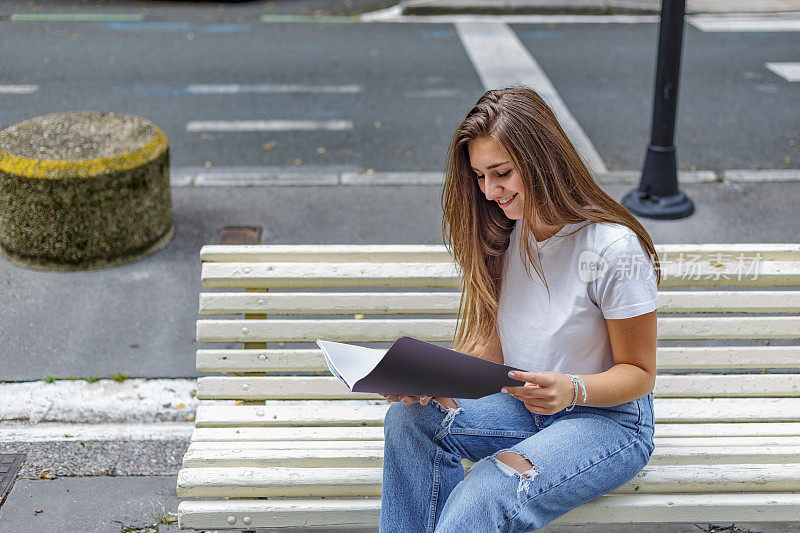 微笑的年轻女子坐在街头长椅上打开一个文件夹