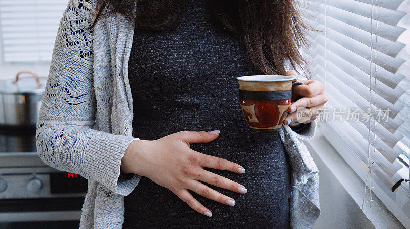 一位孕妇在家里的窗户边喝着咖啡，抚摸着自己的肚子。