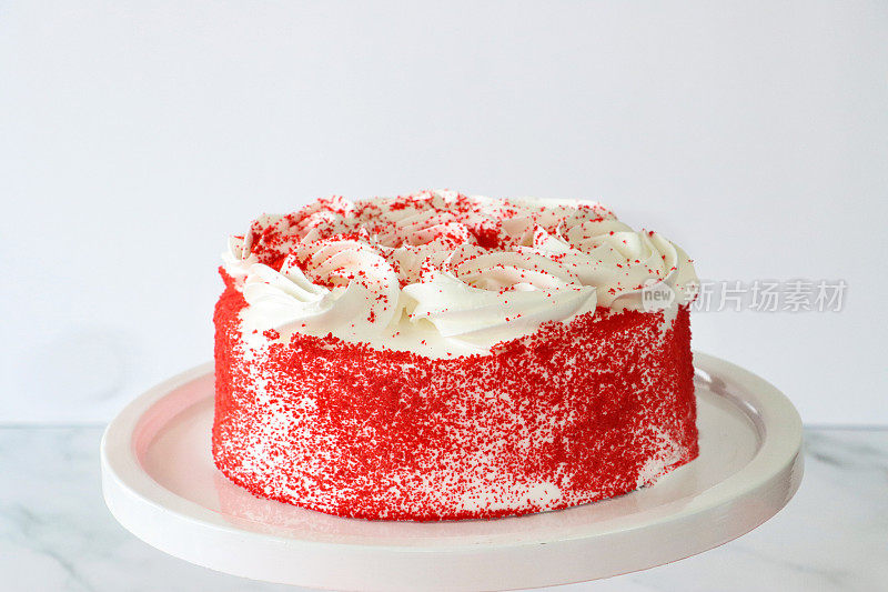 特写图像整个红色天鹅绒蛋糕覆盖黄油奶油，装饰有管道糖衣漩涡和脱水覆盆子粉，在白色的蛋糕架，大理石效果表面，白色背景，重点在前景