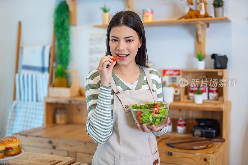 年轻美丽的亚洲女性女士穿着便装和围裙快乐享受吃沙拉与樱桃番茄在她的房子的厨房节食和减肥健康的生活方式