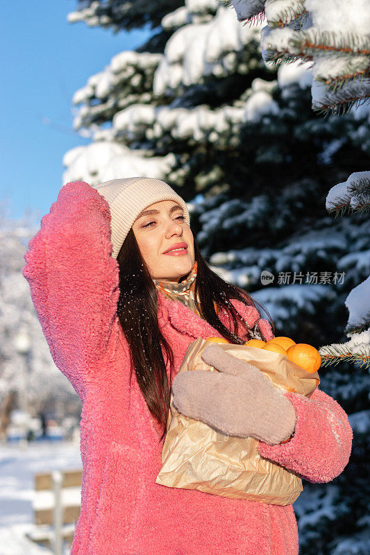 穿着粉红色外套的年轻快乐的女子，手里拿着装着橘子的工艺包，走在白雪纷飞的冬日公园里。节日的气氛中。新年，圣诞节，冬天的概念