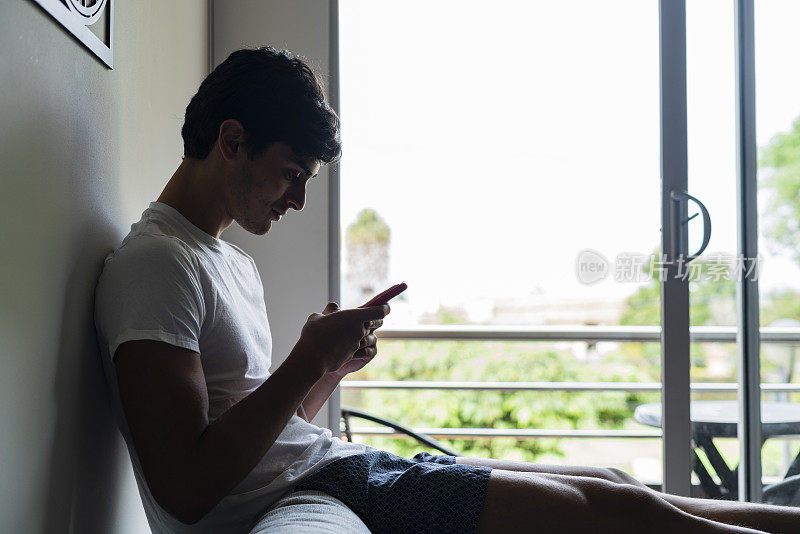 一个年轻的拉丁裔男子坐在他家客厅的沙发上，手里拿着手机查看他的银行信息