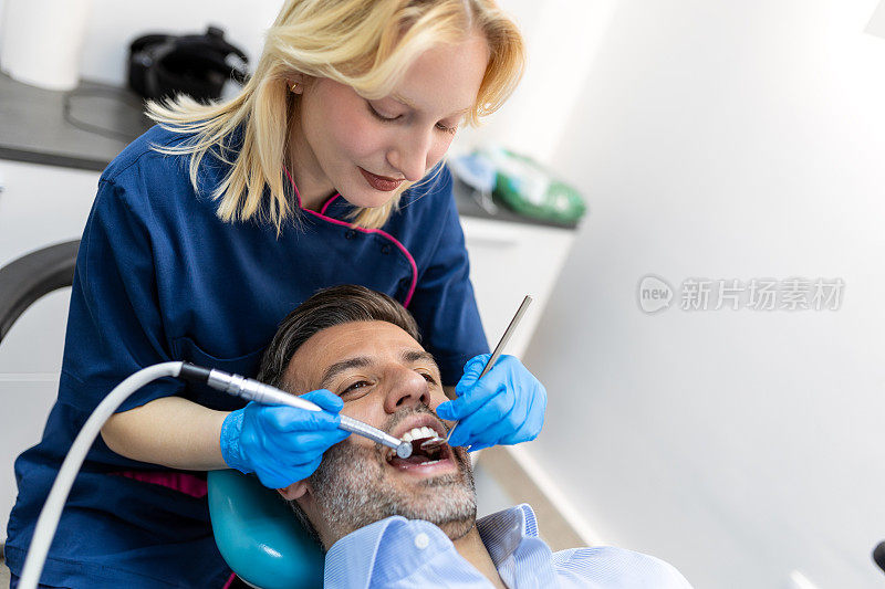 开朗的女牙医拿着牙钻，微笑着靠近病人，女牙医给坐在牙科椅上的男病人钻牙