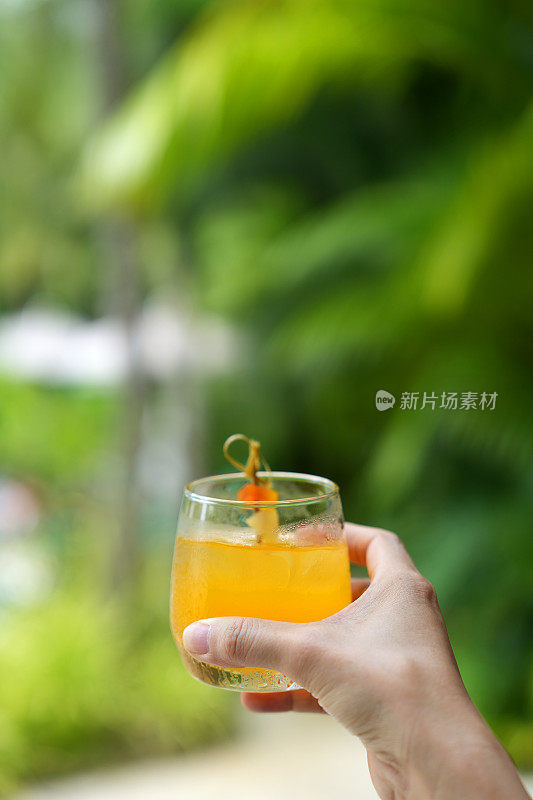 一名年轻女子在欣赏美丽的热带花园和游泳池时，拿着一杯水果潘趣酒。