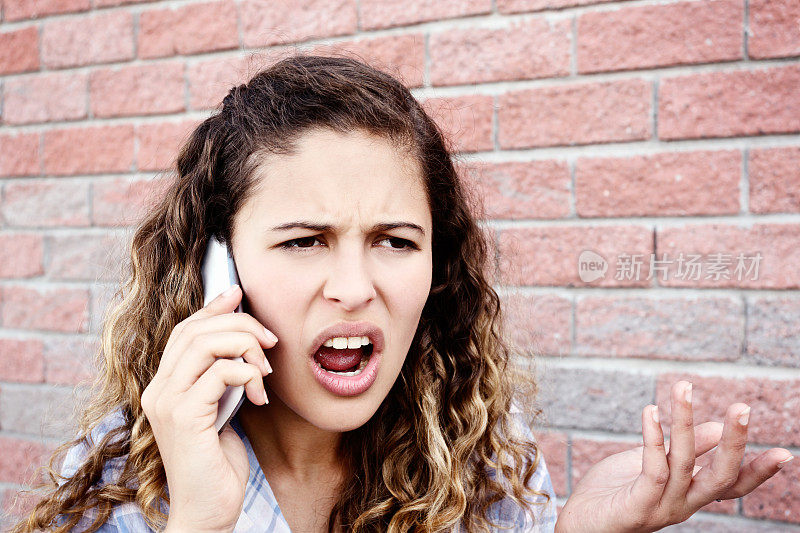 可爱的年轻女子在砖墙边用智能手机愤怒地争吵