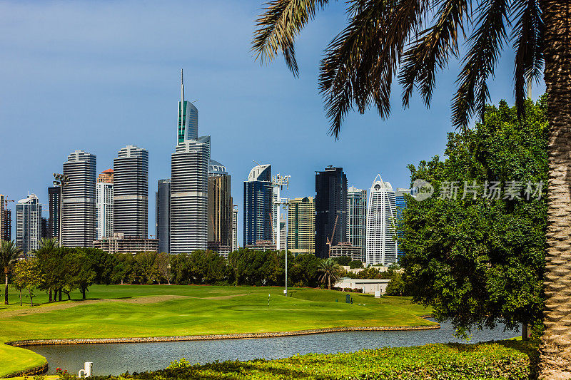 阿拉伯联合酋长国迪拜:高尔夫球道和水的前景;现代摩天楼的JLT在背景中，被一棵椰枣树框在右边-豪华的阿拉伯
