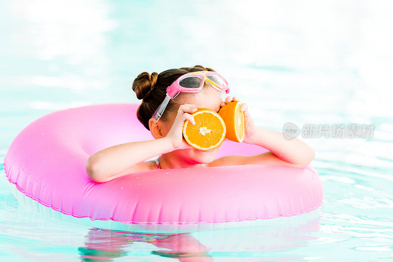 带着充气圈在游泳池游泳的孩子拿着半个橙子在眼睛附近