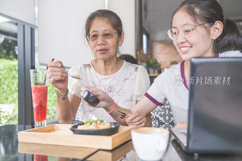 亚洲家庭年轻的青少年和母亲度假吃甜点和喝果汁，使用数字平板电脑和智能手机在咖啡馆放松与在线游戏和在社交网络上的短信