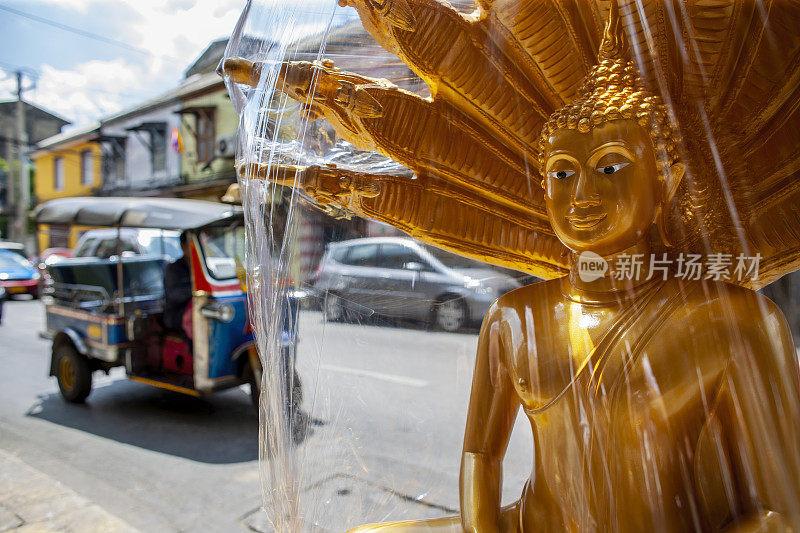 曼谷一家街头商店外出售用塑料包裹的黄金佛像，背景是一辆泰国嘟嘟车。