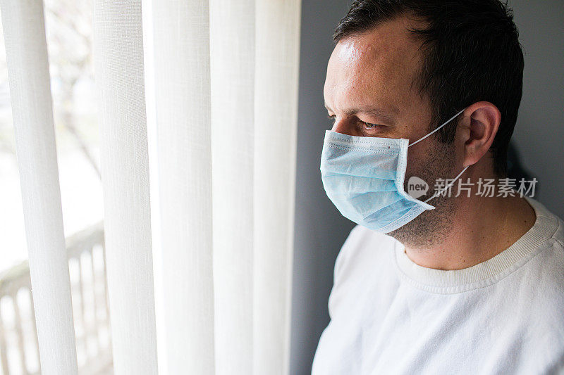 一个戴医用面具的孤独男人透过窗户看。居家隔离自我隔离