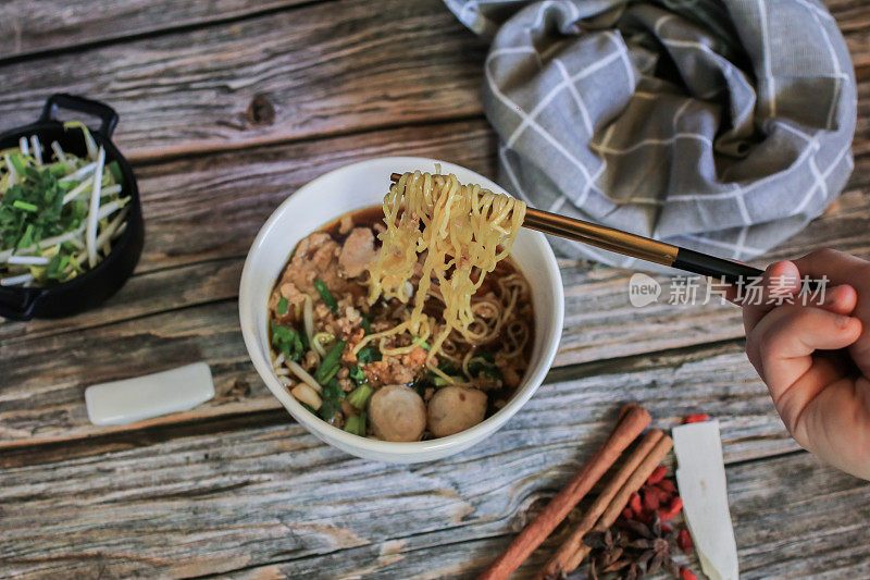 手用筷子夹起美味的鸡蛋面条猪肉和蔬菜在五种香料黑汤在木制桌子上俯视图-亚洲美食风格。