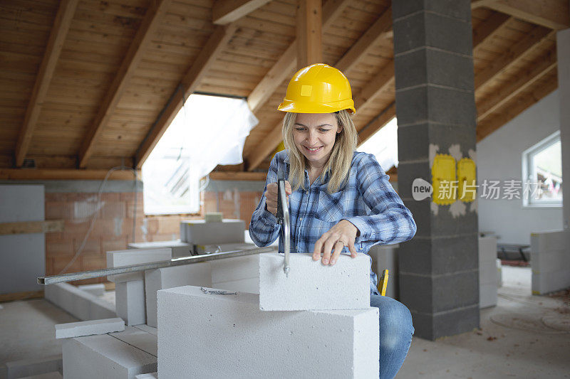 年轻漂亮的女砖匠戴着黄色的安全帽，正在房子里的建筑工地上锯砖