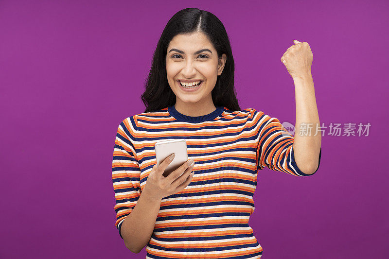 一个年轻开朗的女人的肖像，拿着手机，举起拳头孤立地站在紫色的背景