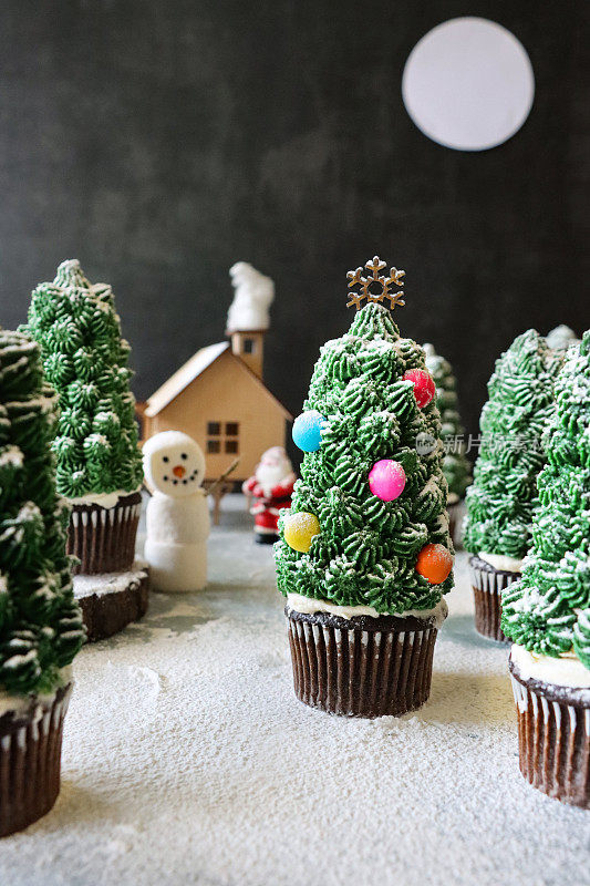 这是一组自制的，圣诞树设计的巧克力纸杯蛋糕，放在纸杯蛋糕盒里，展示在下雪的夜晚针叶林场景中，有圣诞老人的模型房子，棉花糖雪人和满月