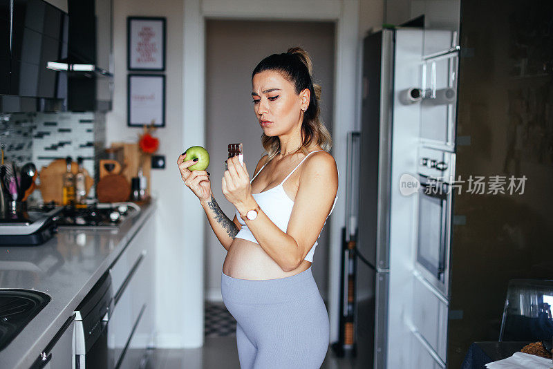年轻的孕妇在厨房里，手里拿着苹果和巧克力。健康食品的概念。