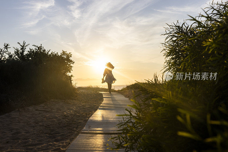 梦幻般的女人与夏装走在日落的海滩路径，撒丁岛，意大利。