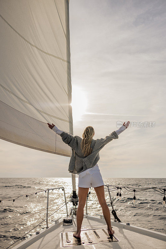 长发女子站在海上帆船的前甲板上，伸开双臂欣赏夕阳