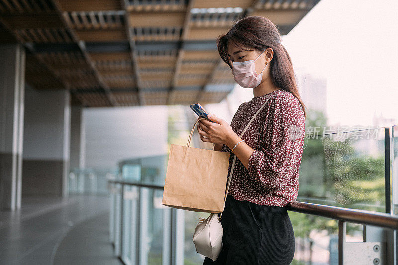 年轻的亚洲女性戴着口罩，拿着购物袋在街上用她的智能手机