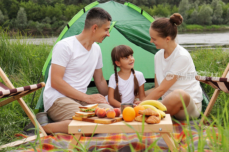 户外拍摄的幸福家庭坐在帐篷附近的地面上，一起野餐，享受美味的水果和三明治，谈论和度过快乐的周末。