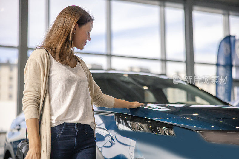 一名女子站在汽车经销商的室内寻找新车，脸上带着微笑摸着一辆新SUV的发动机罩