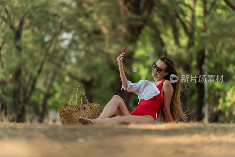 美丽的白人女性性感的女人在红色泳装比基尼戴着太阳镜自拍使用她的智能手机快乐和放松在海边的树林